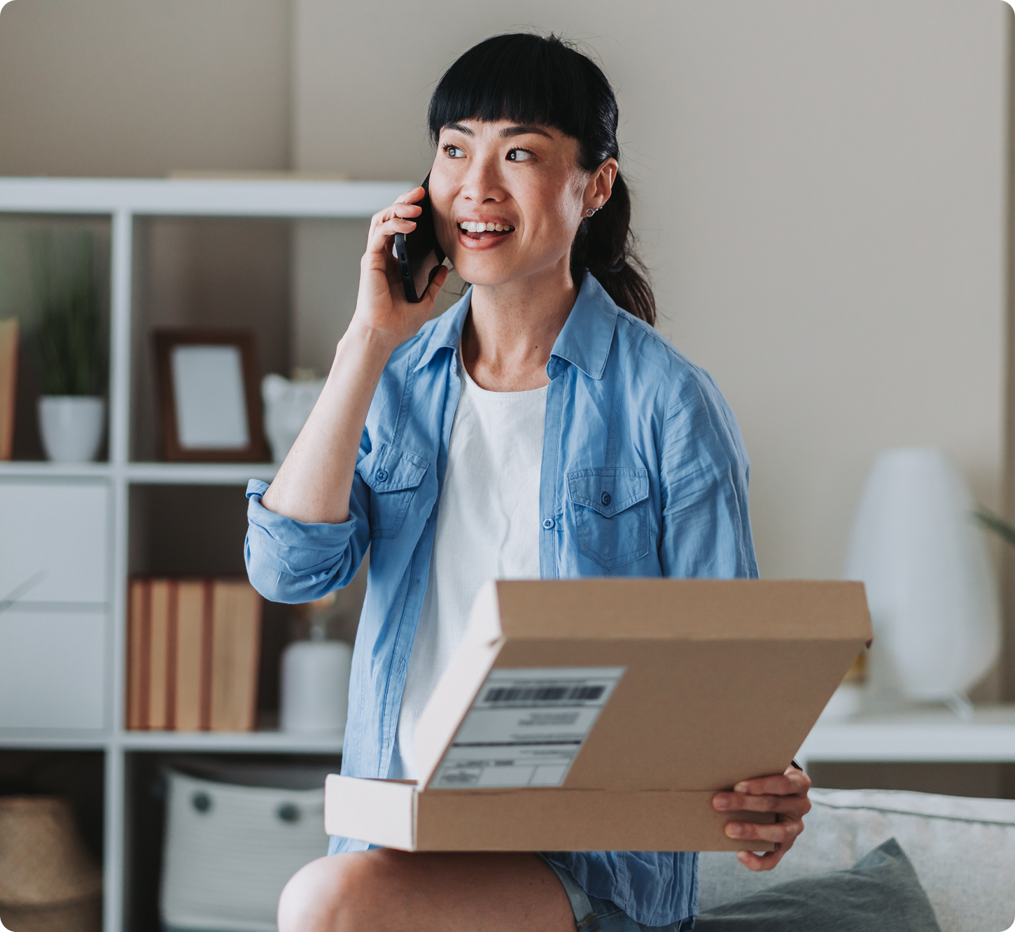 A woman smiles while talking on the phone, holding an open cardboard box in a cozy living room. She wears a blue shirt and white top, with shelves in the background.