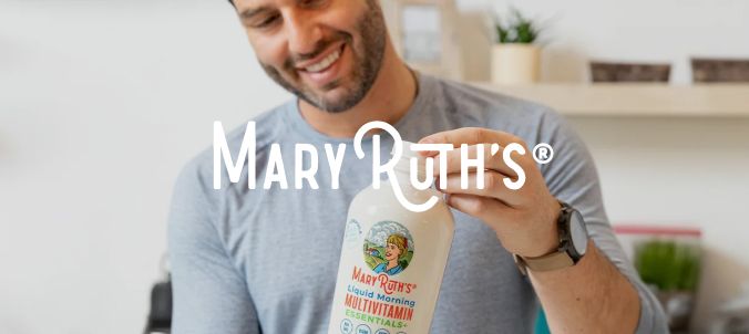 A smiling man holds a MaryRuth's multivitamin bottle in a modern kitchen, conveying health and wellness. Shelving and kitchenware are in the background.