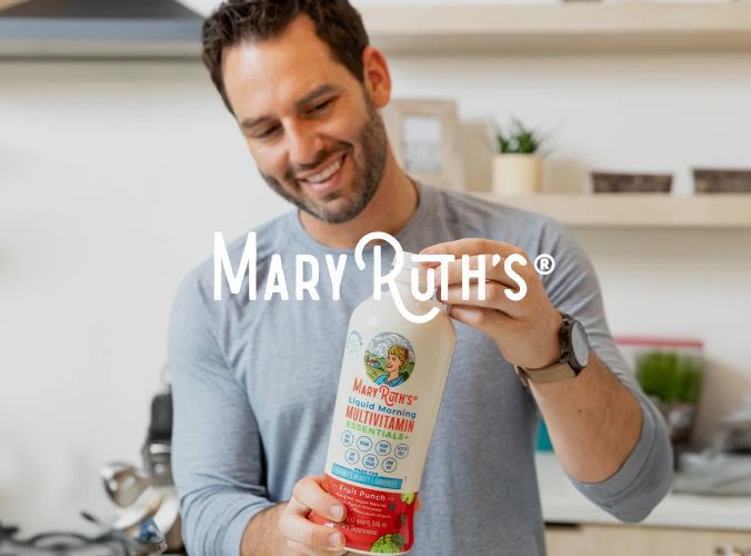 A smiling man holds a MaryRuth's multivitamin bottle in a modern kitchen, conveying health and wellness. Shelving and kitchenware are in the background.