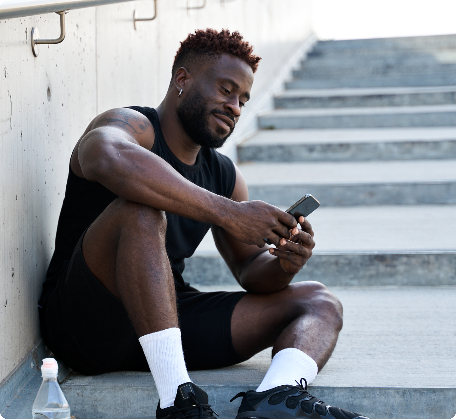 Man in athletic wear sits on outdoor steps, smiling while looking at his smartphone. A water bottle is beside him, conveying a relaxed, post-workout vibe.