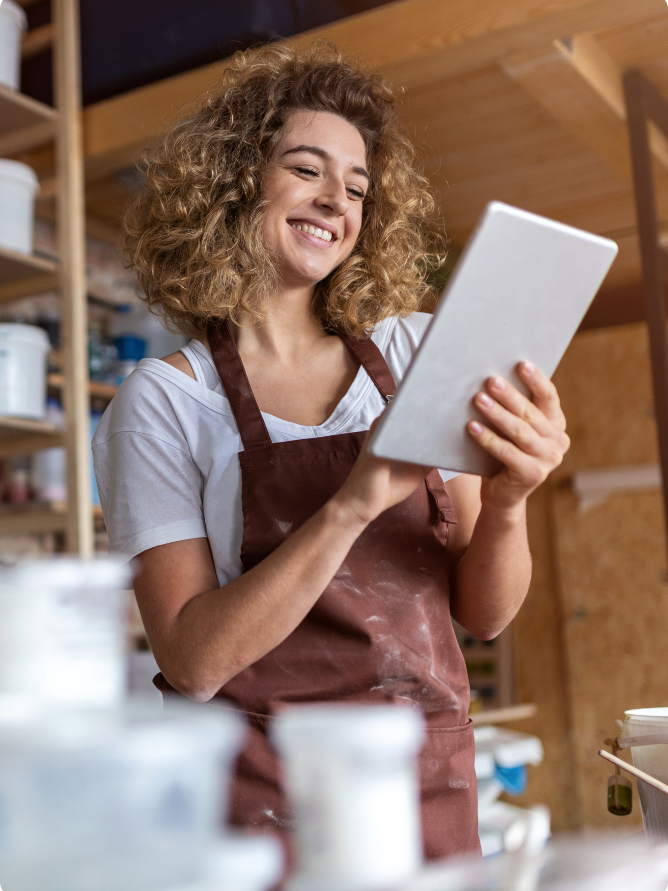 Smiling woman with curly hair in a brown apron holding a tablet in a pottery studio. Shelves with containers are visible. The mood is happy and creative.