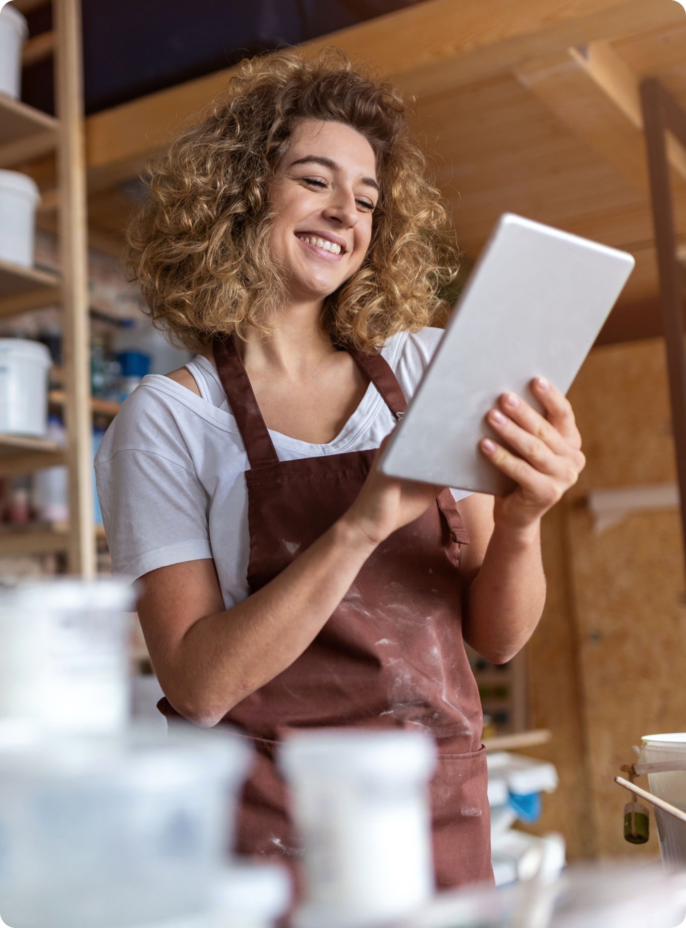 Smiling woman with curly hair in a brown apron holding a tablet in a pottery studio. Shelves with containers are visible. The mood is happy and creative.