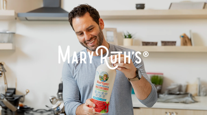 A smiling man holds a large bottle of Mary Ruth's Liquid Morning Multivitamin in a modern kitchen. The setting is bright and inviting, suggesting health and wellness.