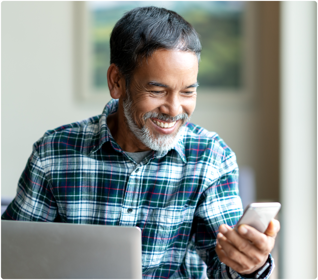 Man with a gray beard in a plaid shirt smiles while looking at a smartphone. He sits at a table with a laptop, conveying a cheerful mood.