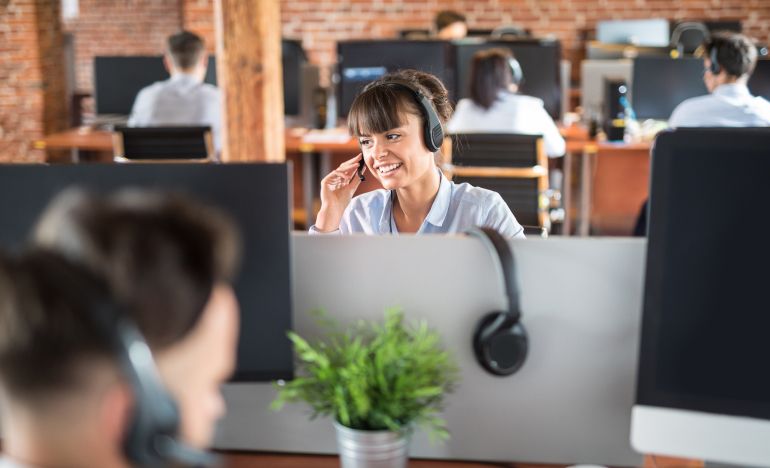 A woman in bangs wearing a phone headset sitting in a customer service office and smiling