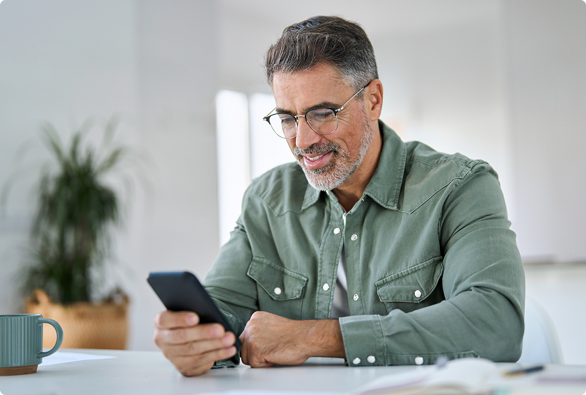 A middle-aged man with glasses and a beard smiles while looking at his smartphone. He wears a green shirt and sits at a white table with a mug.