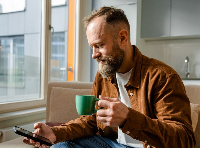 A bearded man in casual attire sips from a green mug, browsing on his phone