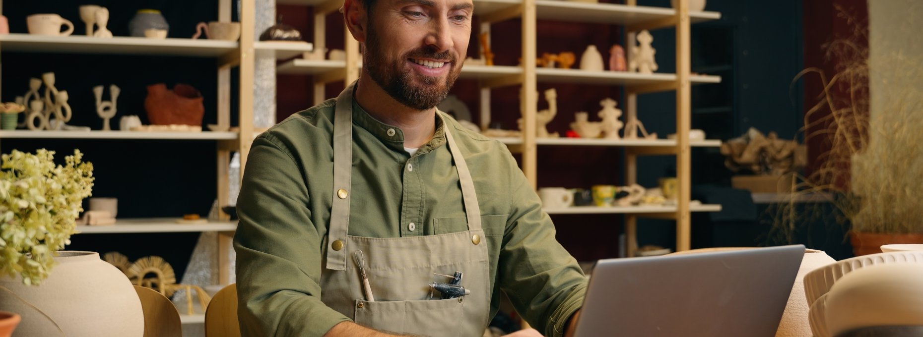 Smiling small-business owner in a canvas apron works on a laptop in his ceramics shop, with shelves of pottery and vases behind him