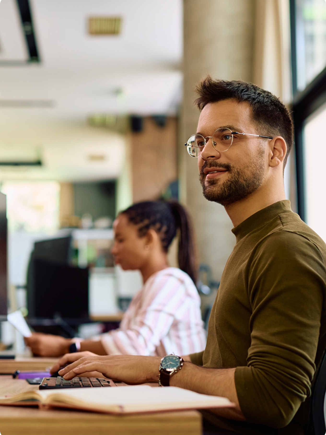 A man with glasses, wearing a green sweater, smiles while working on a computer in a bright office. A woman in a striped shirt works behind him.