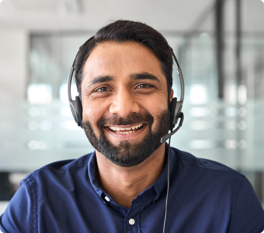 Smiling man with a beard wearing a headset in an office setting. He exudes a friendly, professional demeanor. The background includes blurred glass panels.