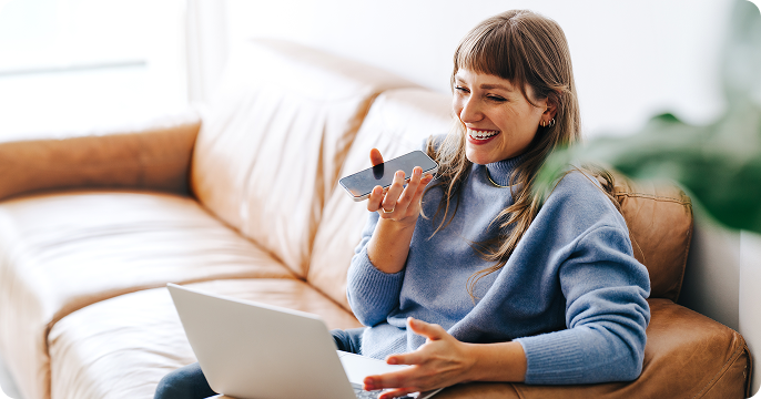 Smiling woman in a blue sweater, sitting on a couch, using voice command on a smartphone, with an open laptop beside her, conveying a cheerful mood.