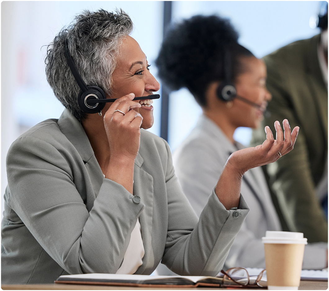 An older woman in a headset, smiling while on a call, sits at a desk with papers and a coffee cup. Colleagues in the background appear engaged.