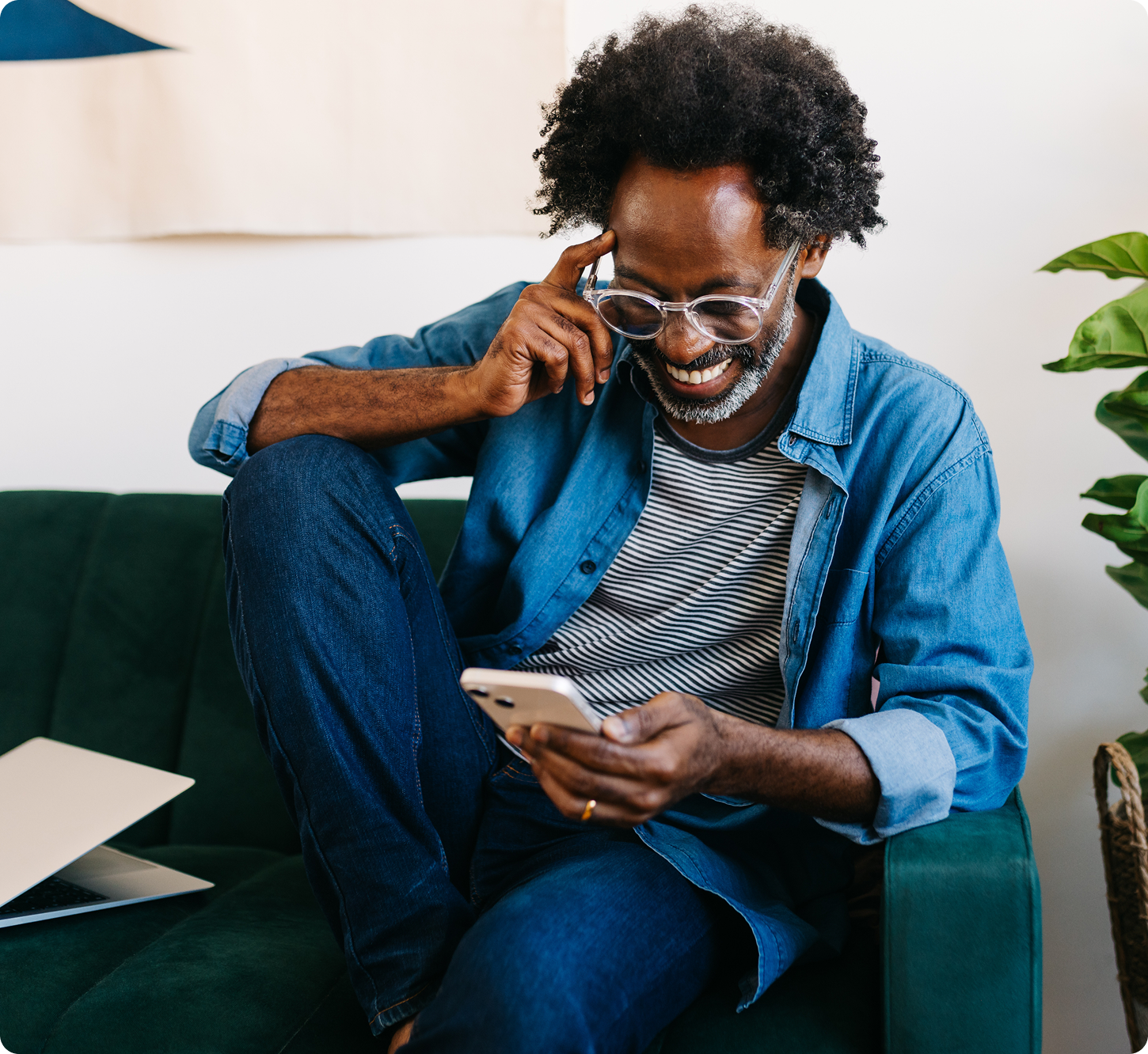 A man with curly hair and glasses smiles while looking at his phone. He's wearing a denim shirt and striped shirt, sitting on a green couch.