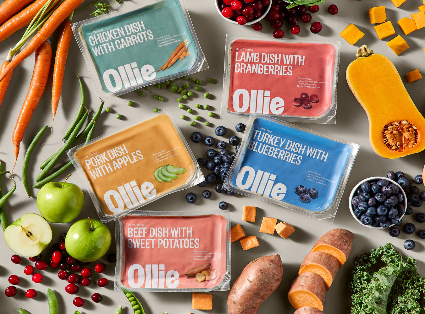 Packages of Ollie pet food with flavors like chicken, lamb, pork, and beef are surrounded by fresh ingredients like carrots, cranberries, apples, and sweet potatoes.