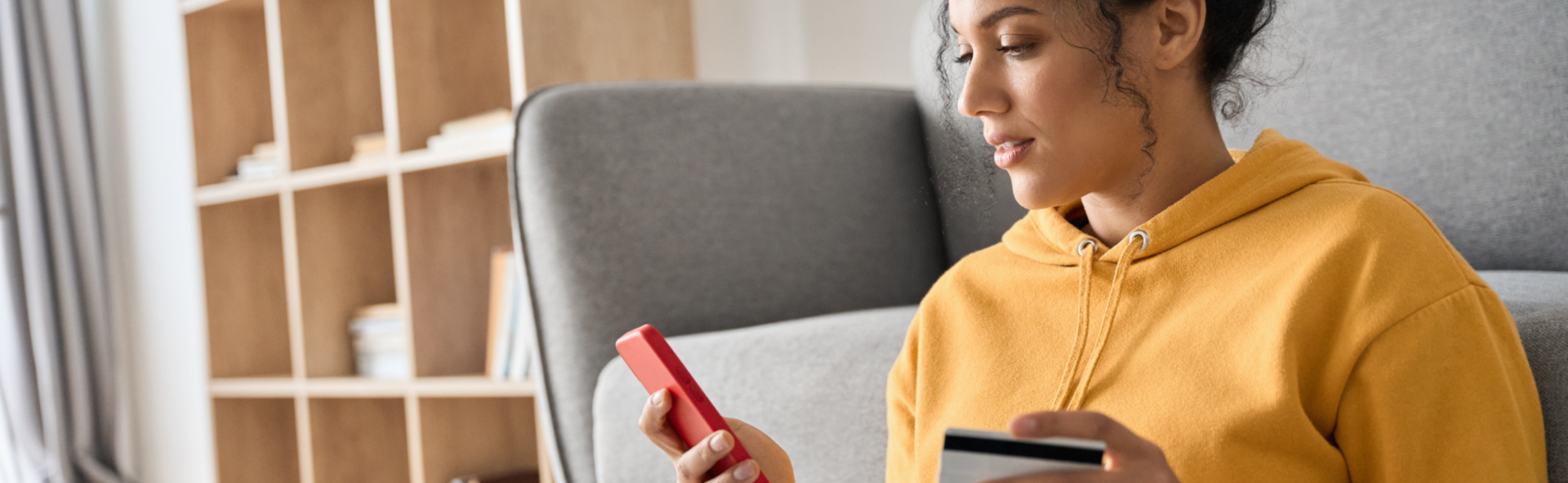 A woman in a yellow sweatshirt in her living room buying something online with her red smartphone, a credit card in her hand