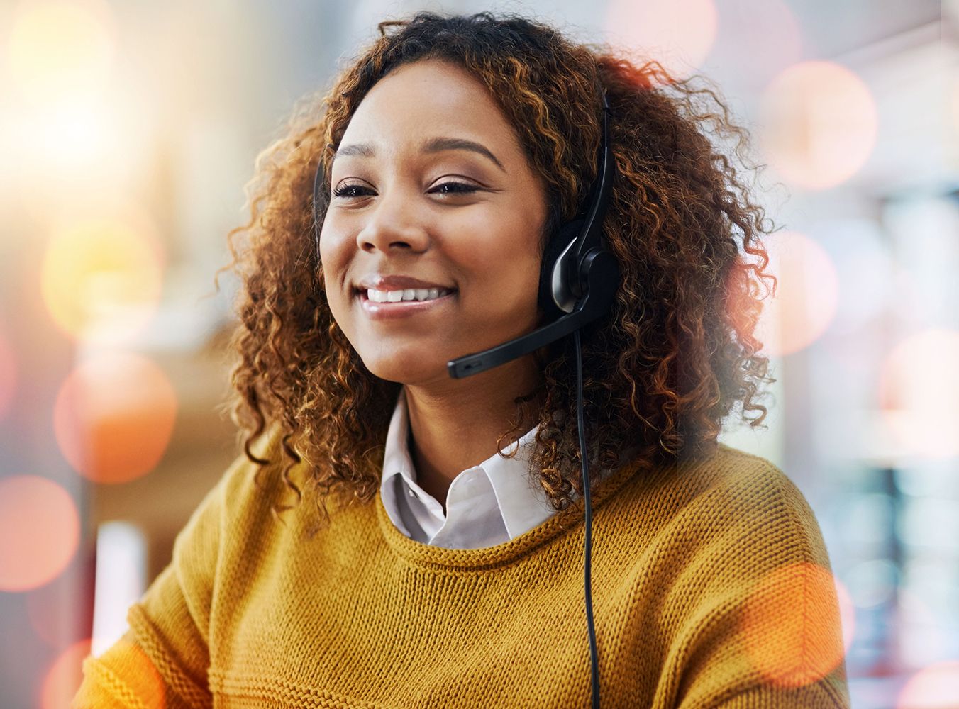 Smiling woman wearing a headset, sitting in a well-lit office. Her curly hair frames her face, and she's dressed in a mustard sweater, creating a warm atmosphere.