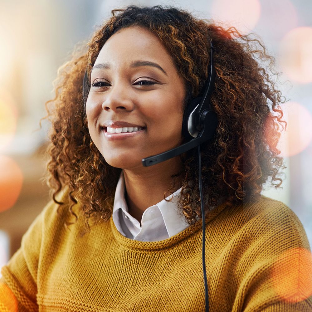 Smiling woman wearing a headset, sitting in a well-lit office. Her curly hair frames her face, and she's dressed in a mustard sweater, creating a warm atmosphere.