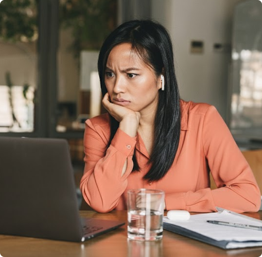 A woman in an orange blouse, looking frustrated or deep in thought, sits at a table with a laptop, glass of water, and papers in a bright office space.