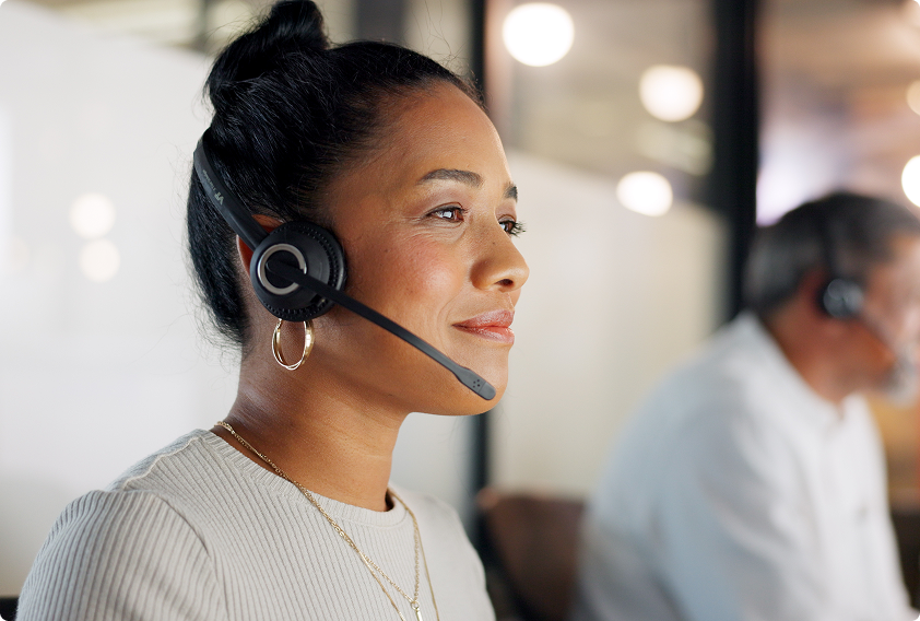 Woman with a headset smiles at work, suggesting a customer service setting. Blurred background with another person, conveying a busy yet calm atmosphere.