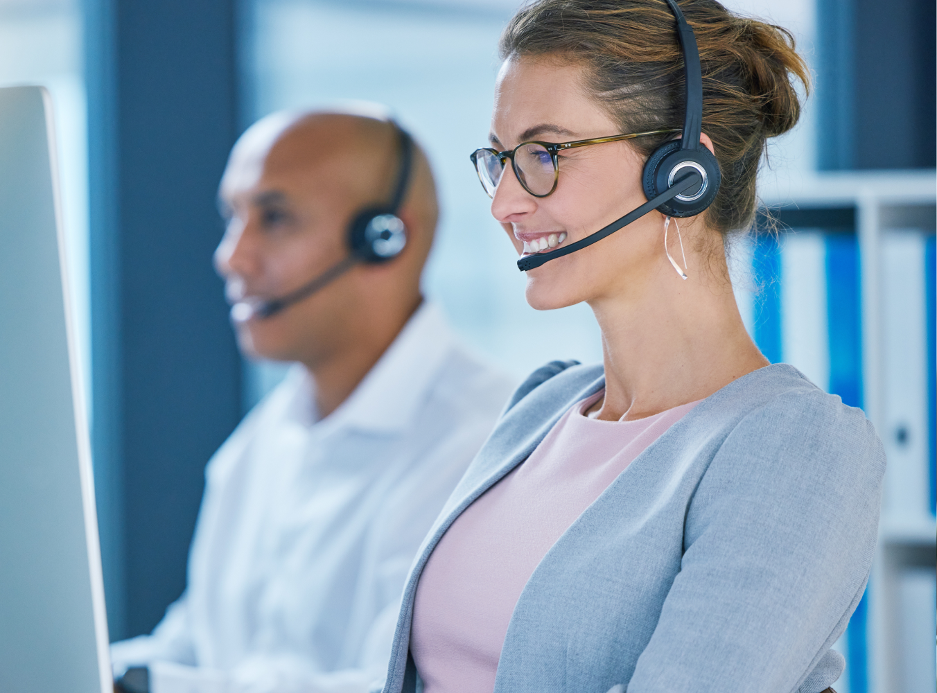 A woman in glasses and a headset smiles, working at a computer. In the background, a man with a headset sits at another desk. The scene is professional and focused.