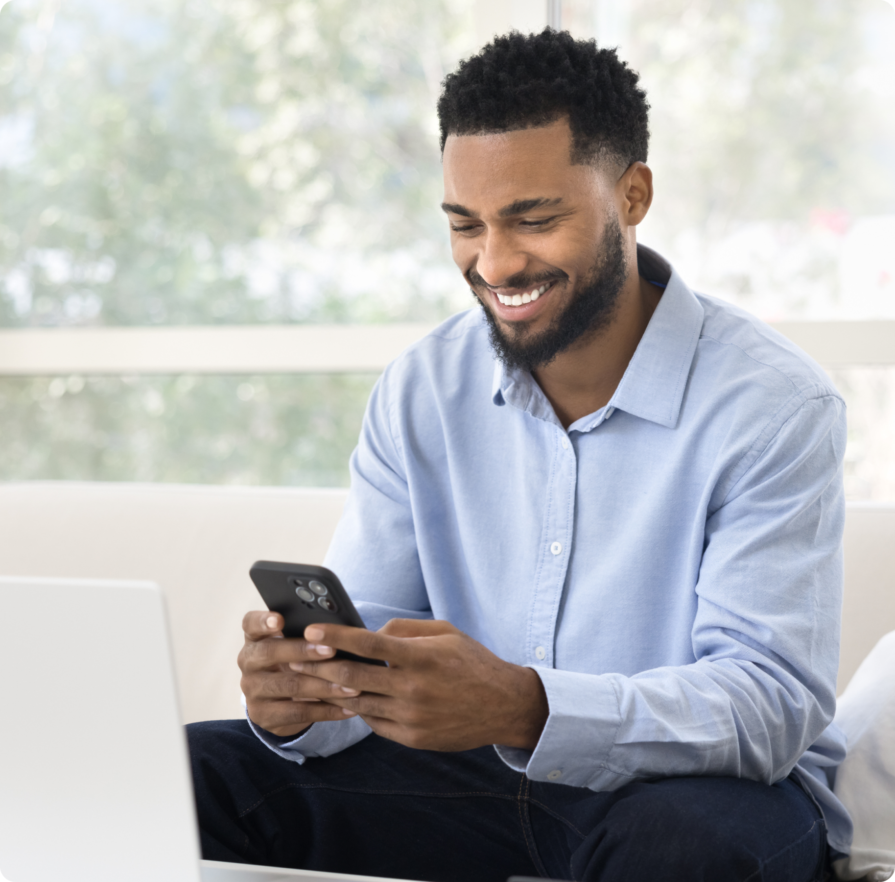Smiling man in a light blue shirt uses a smartphone indoors, seated comfortably with a blurred nature background. He appears relaxed and happy.