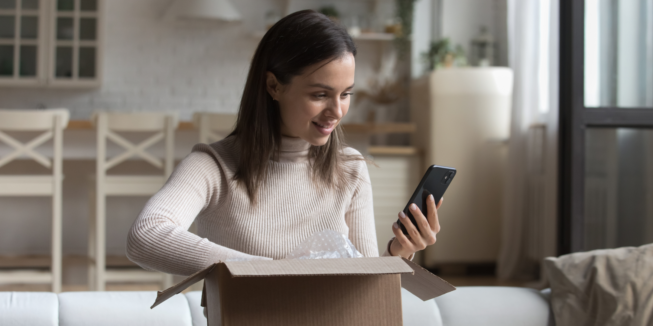 Woman smiling as she opens a package, holding a smartphone in one hand in a cozy living room. The scene conveys excitement and anticipation.