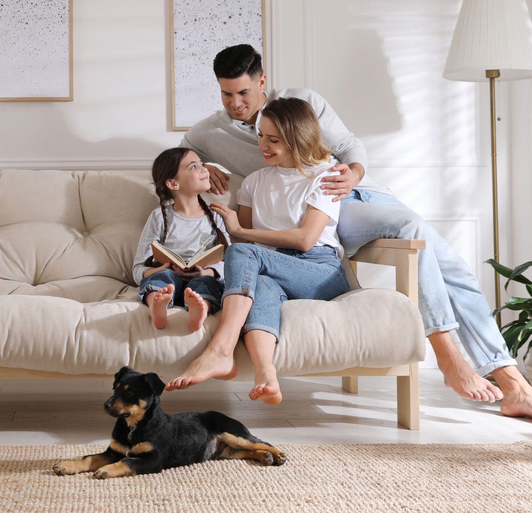 Family sitting on a beige sofa, with a child holding a book. Parents in casual clothes, smiling warmly. Black and tan dog resting on the rug. Cozy atmosphere.