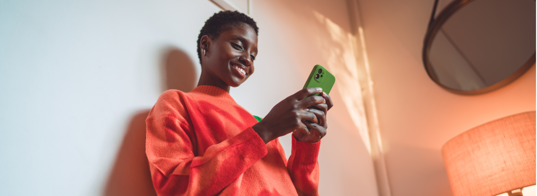 Smiling woman in an orange sweater leans against a wall at home, shopping on her phone