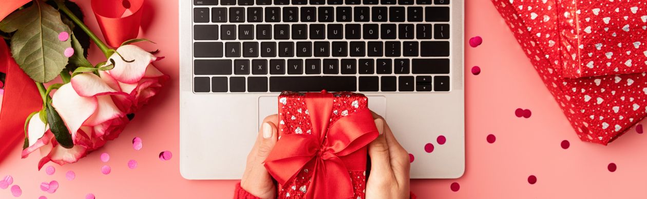 Someone holding a valentine's day gift over a laptop on a desk scattered with valentine's day decorations