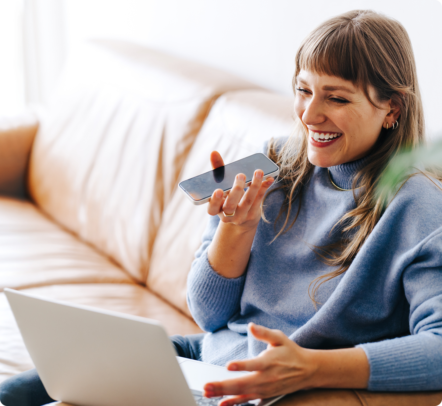 Smiling woman in a blue sweater, sitting on a couch, using voice command on a smartphone, with an open laptop beside her, conveying a cheerful mood.