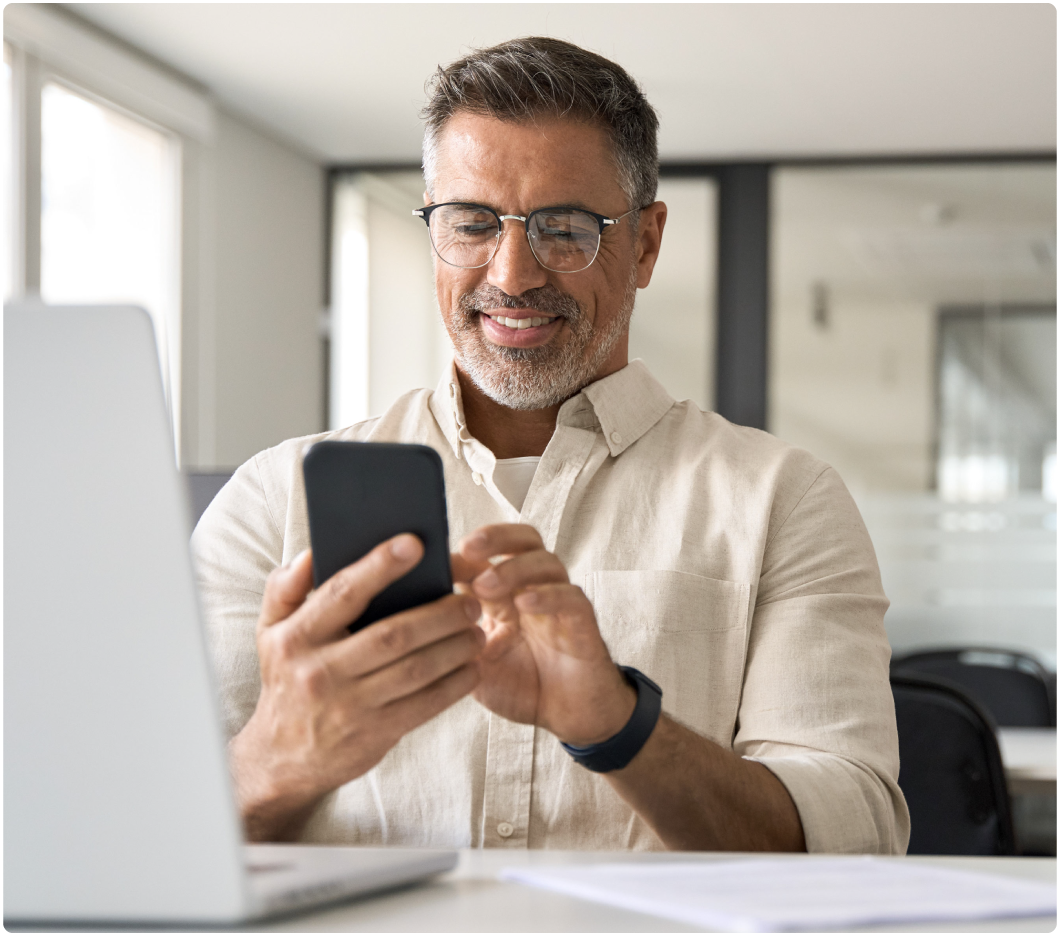 Man with glasses in a light shirt smiles while typing on a smartphone at a modern office desk, with a laptop and documents nearby, conveying focus.
