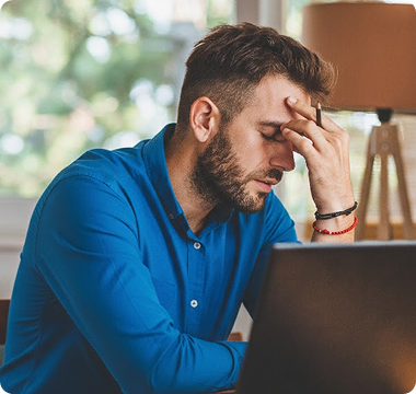 A man in a blue shirt sits at a desk with a laptop, looking stressed, holding his forehead. The room has soft lighting and a blurred view outside.