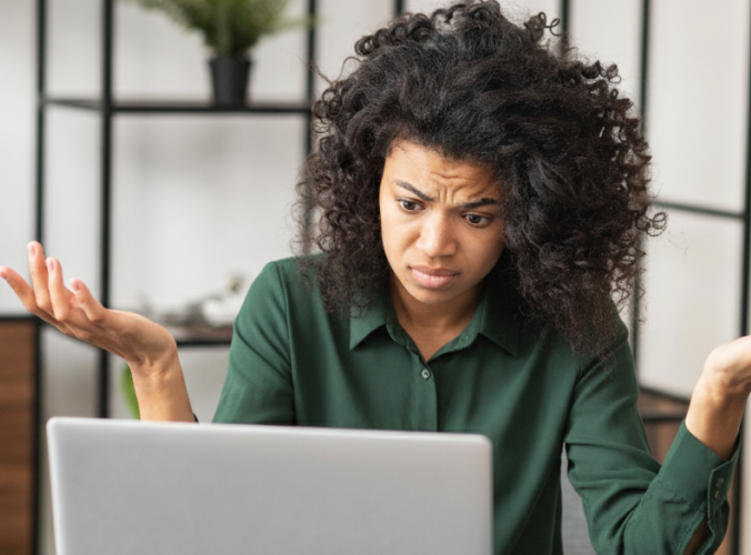 Woman in green shirt looks puzzled at laptop, raising hands in confusion. Background has shelves with a plant. Emotions convey frustration and bewilderment.