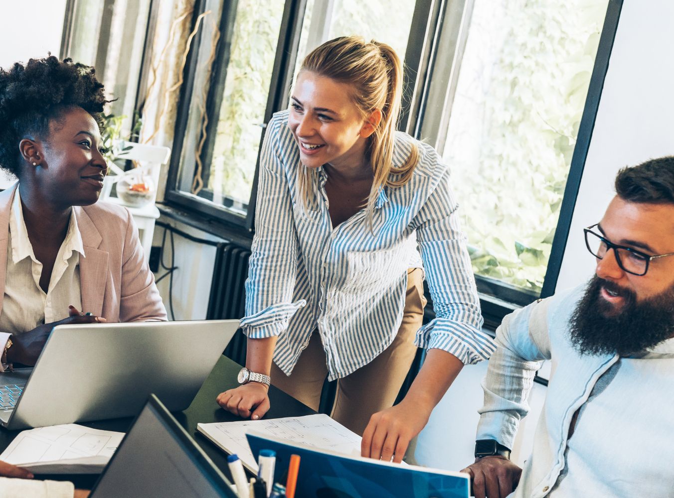 A diverse group of three colleagues in casual attire is collaborating around a table, smiling and engaged, surrounded by laptops and papers in a well-lit office.