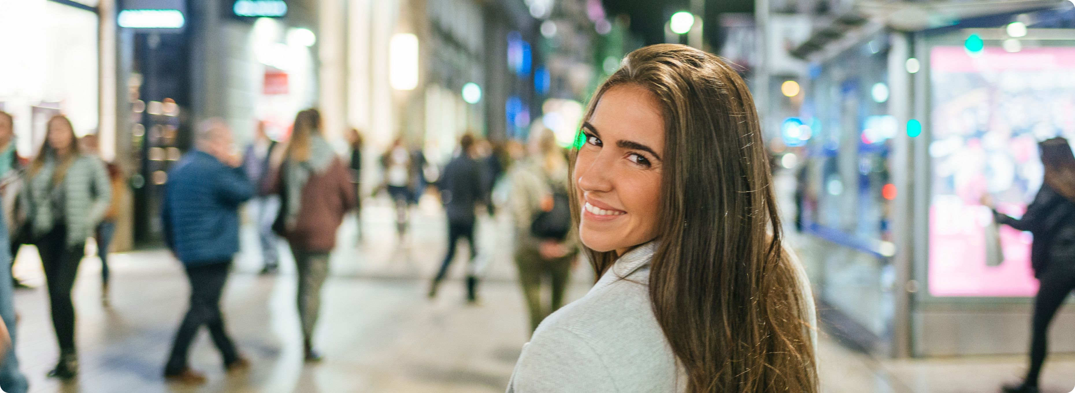 Smiling woman with long brown hair looks back at the camera while walking in a lively, brightly illuminated city street filled with blurred pedestrians.