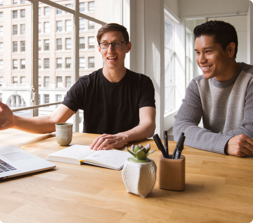 Two men sit at a wooden table in a bright room, smiling and engaged in discussion. A laptop, notebook, and plants are on the table, conveying a collaborative and positive atmosphere.