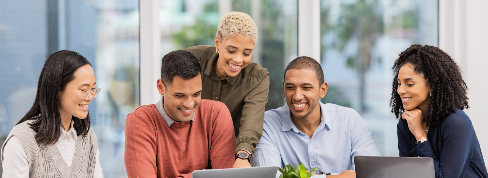 Team of five smiling coworkers gathered around a laptop, planning together at a shared desk