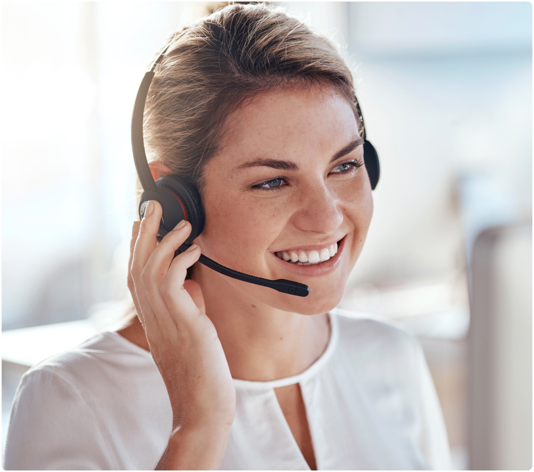 A smiling woman in a white blouse wears a headset in a bright office, suggesting a positive and professional customer service environment.
