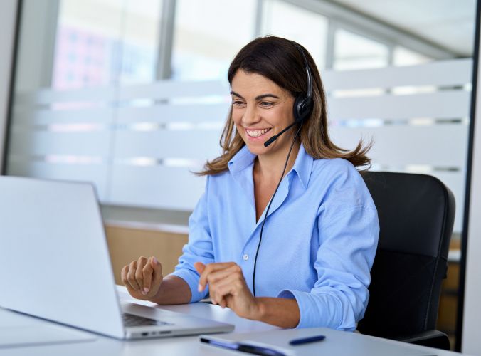 Woman wearing a headset smiles while engaging in a video call on a laptop. She is seated in an office setting, conveying a professional and positive tone.