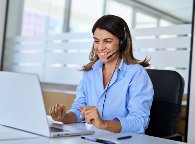 Woman wearing a headset smiles while engaging in a video call on a laptop. She is seated in an office setting, conveying a professional and positive tone.