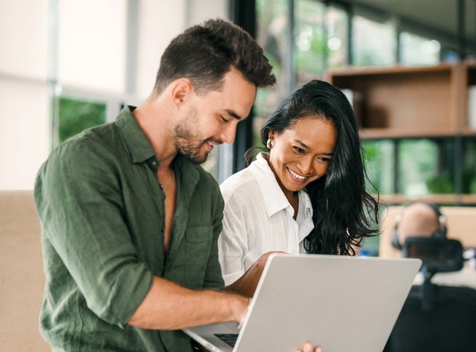 A smiling man and woman collaborate on a laptop in a modern office, with large windows in the background, conveying teamwork and positivity.