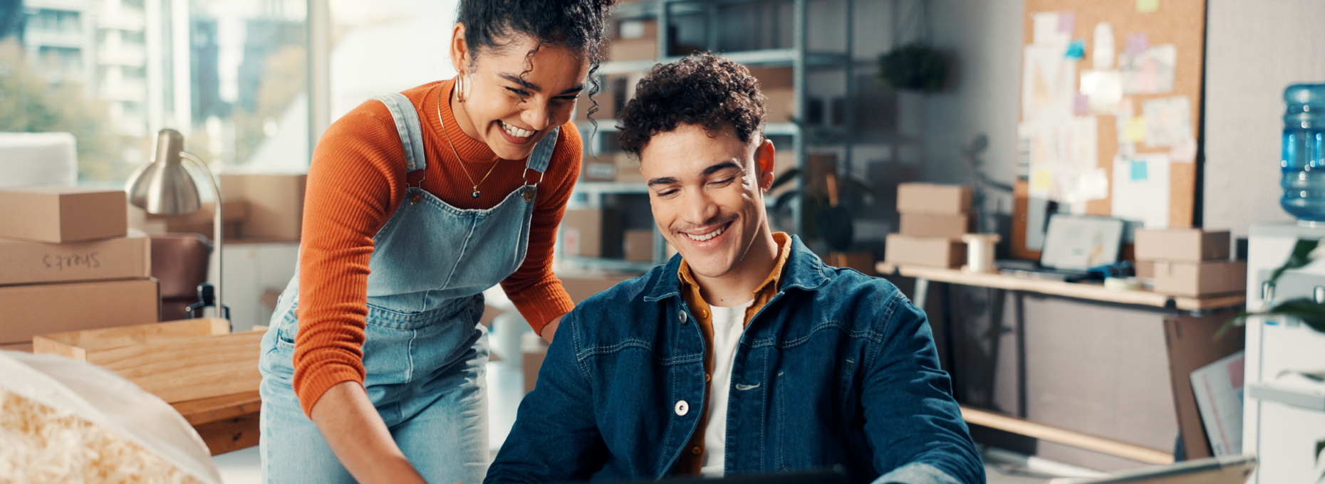 Two coworkers smiling while they review something on a laptop together in a small ecommerce stockroom with shelves and shipping boxes