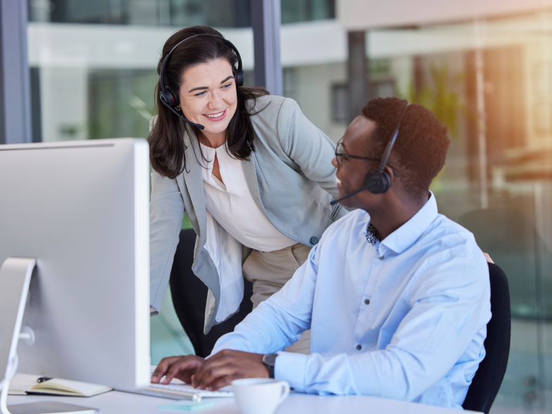 Smiling customer support agent in a headset leaning over to help a colleague at his computer in a bright office