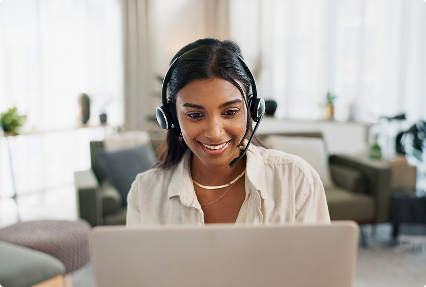 A woman with headphones smiles while looking at a laptop in a cozy living room. Bright, welcoming atmosphere with soft chairs and plants in the background.