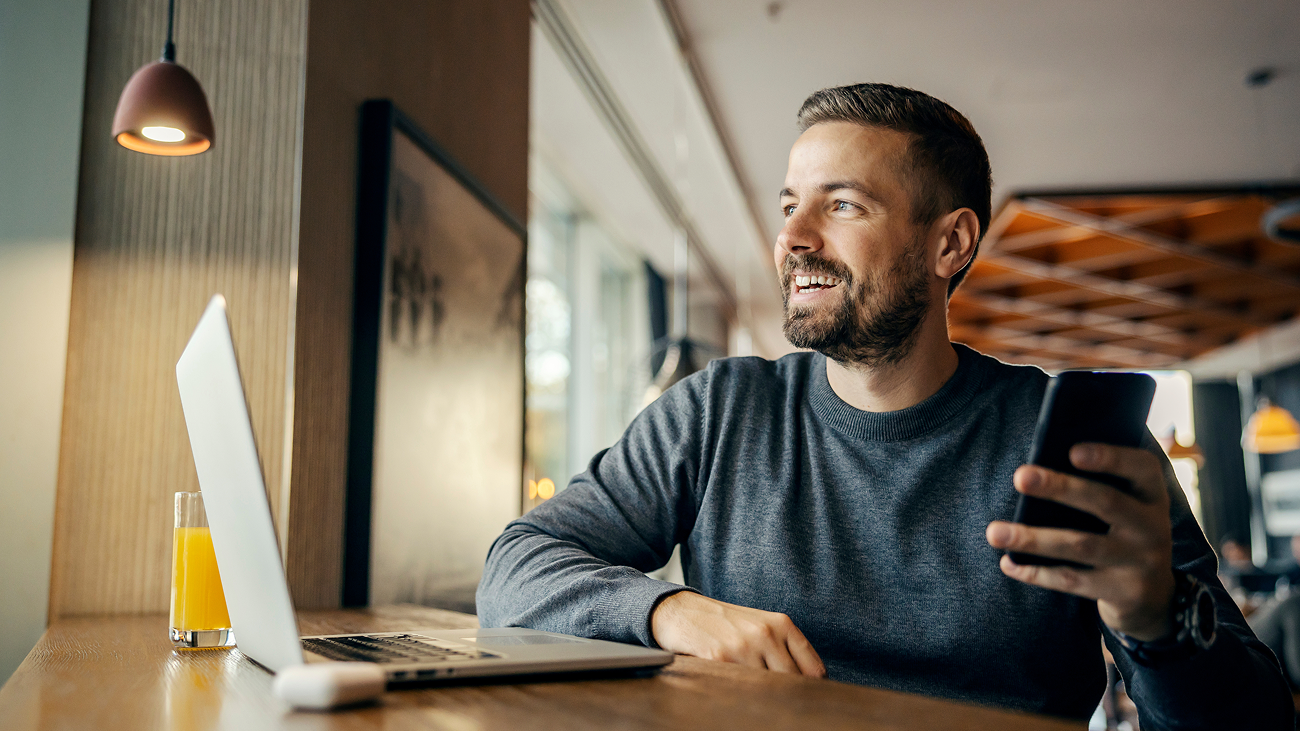 A man sitting in a cafe smiles while looking out a window. He holds a smartphone and sits at a wooden table with a laptop and a glass of orange juice.