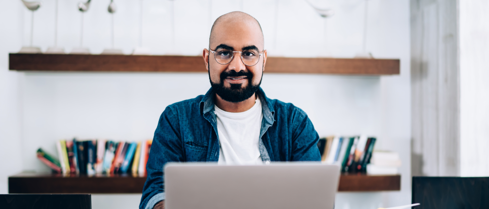 Man in glasses sitting in front of a laptop smiling at the camera
