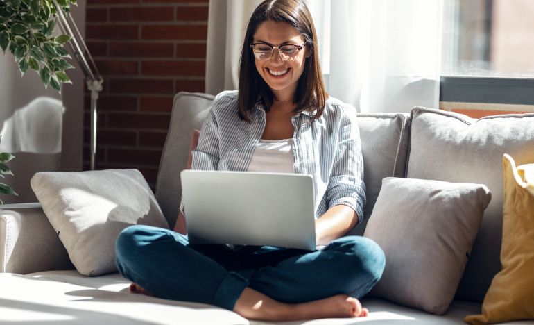 Woman smiling at the laptop in her lap while seated on a gray couch