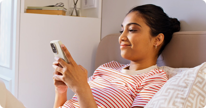 Young woman in a kitchen holds a pink phone, glancing at the screen. She's wearing a blue shirt over a striped top. Sunlight casts soft shadows.