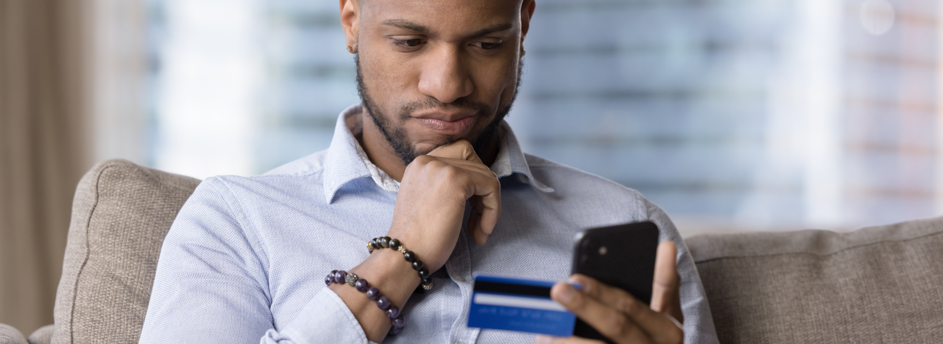 Man on the couch, hand on chin, card in one hand and phone in the other