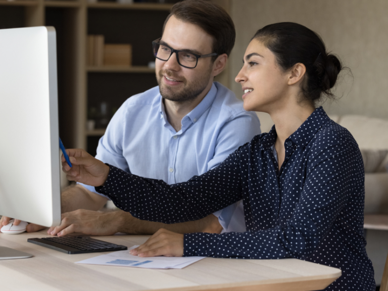 Two colleagues on the computer, one pointing at screen explaining something